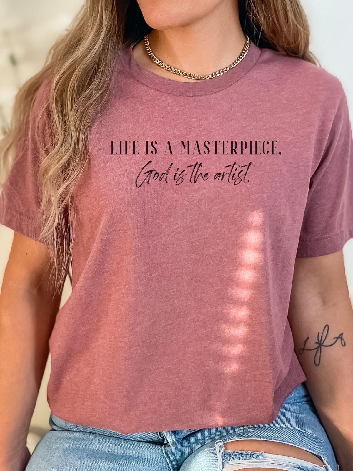 Woman wearing a mauve t-shirt with Life Is A Masterpiece God is the artist text